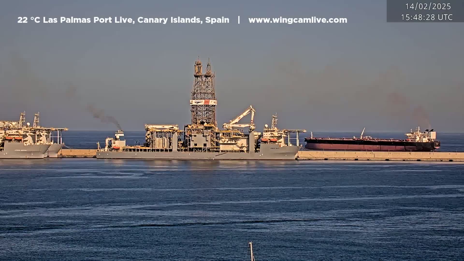 A large drilling rig and several other ships are docked at a port on a calm, sunny day.