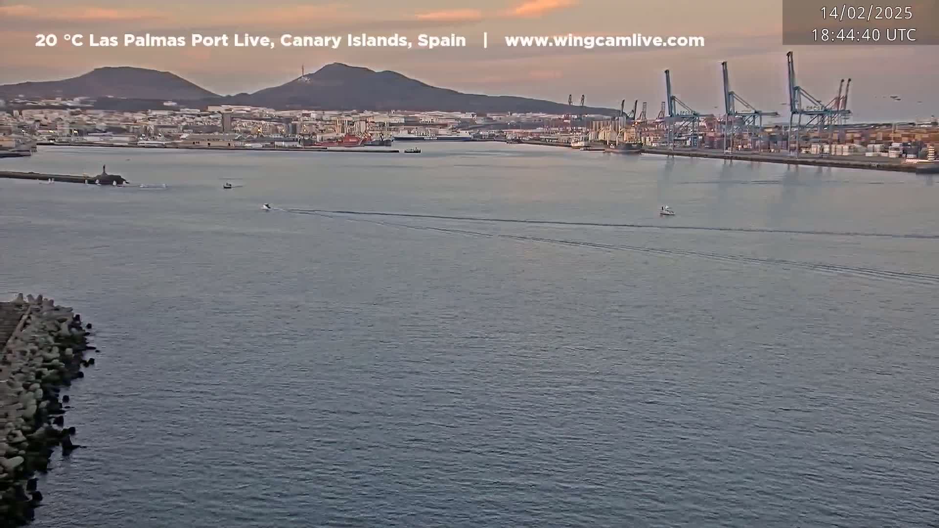 A calm, twilight view of a harbor with several small boats, a city skyline in the background, and large cranes along a dock.