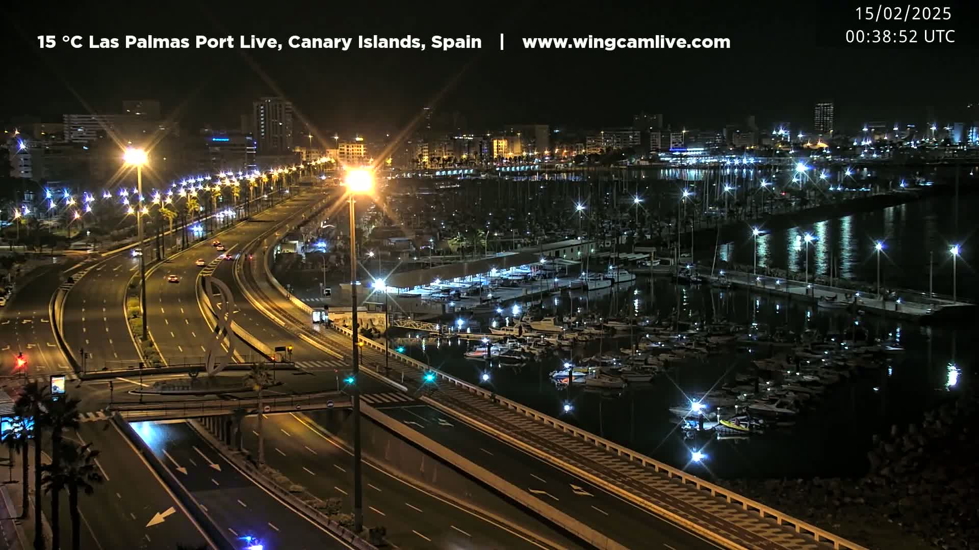 A nighttime, high-angle view shows a marina filled with boats, a highway with sparse traffic, and a city skyline illuminated by streetlights.