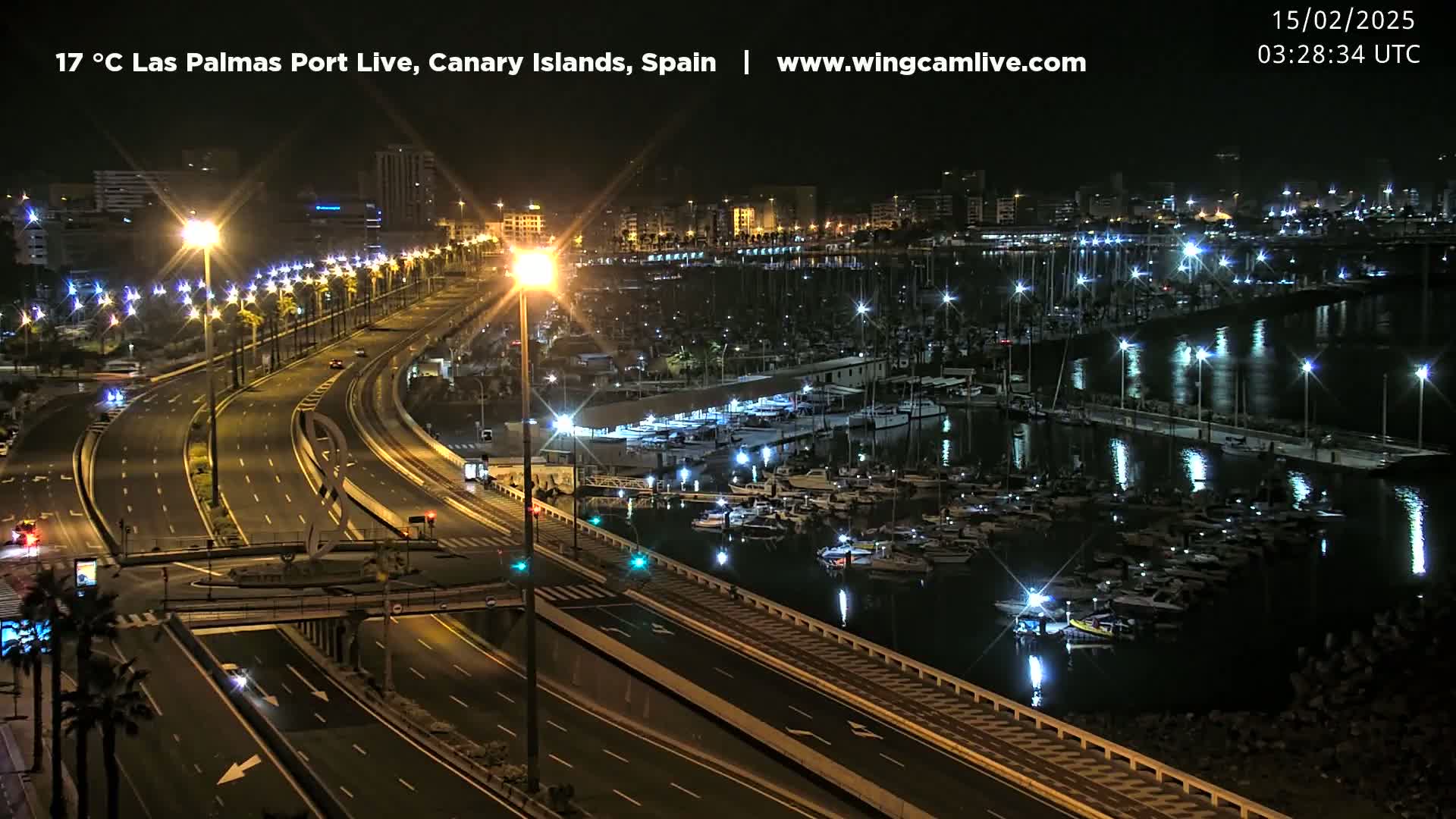 A nighttime, well-lit view of a marina filled with boats and a multi-lane highway beside it.