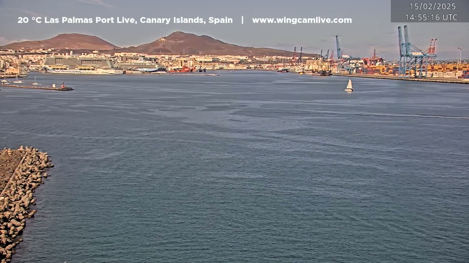 A calm body of water shows a port with docked ships, cargo containers, and cranes under a clear sky.