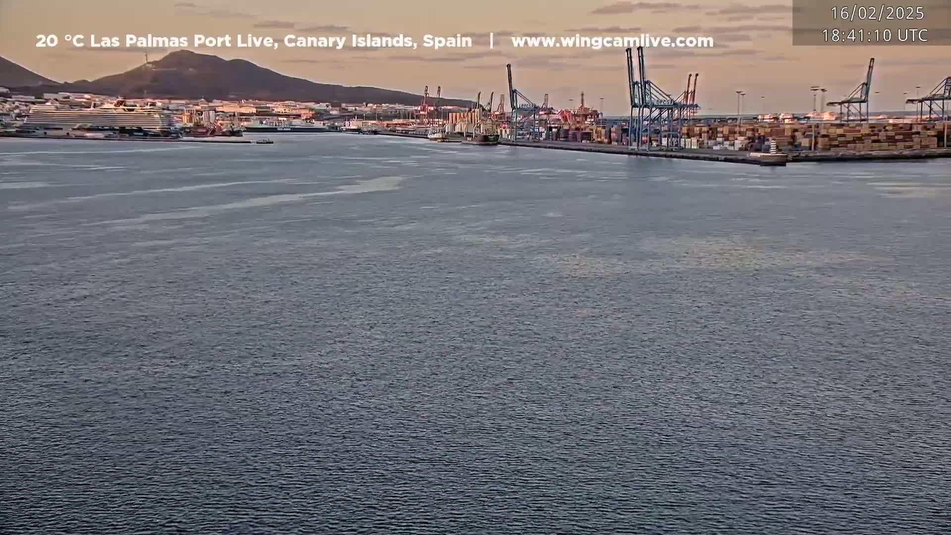A calm body of water shows a port with several cranes and cargo ships under a twilight sky.