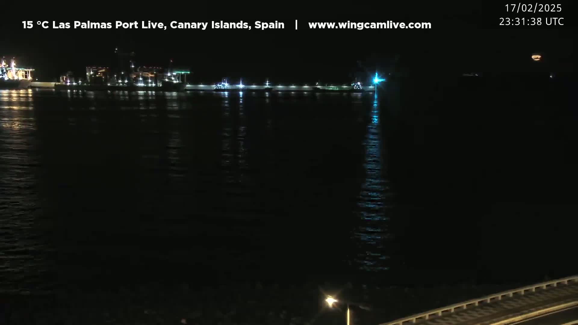 At night, a dark body of water reflects the lights of a distant port, under a clear sky.