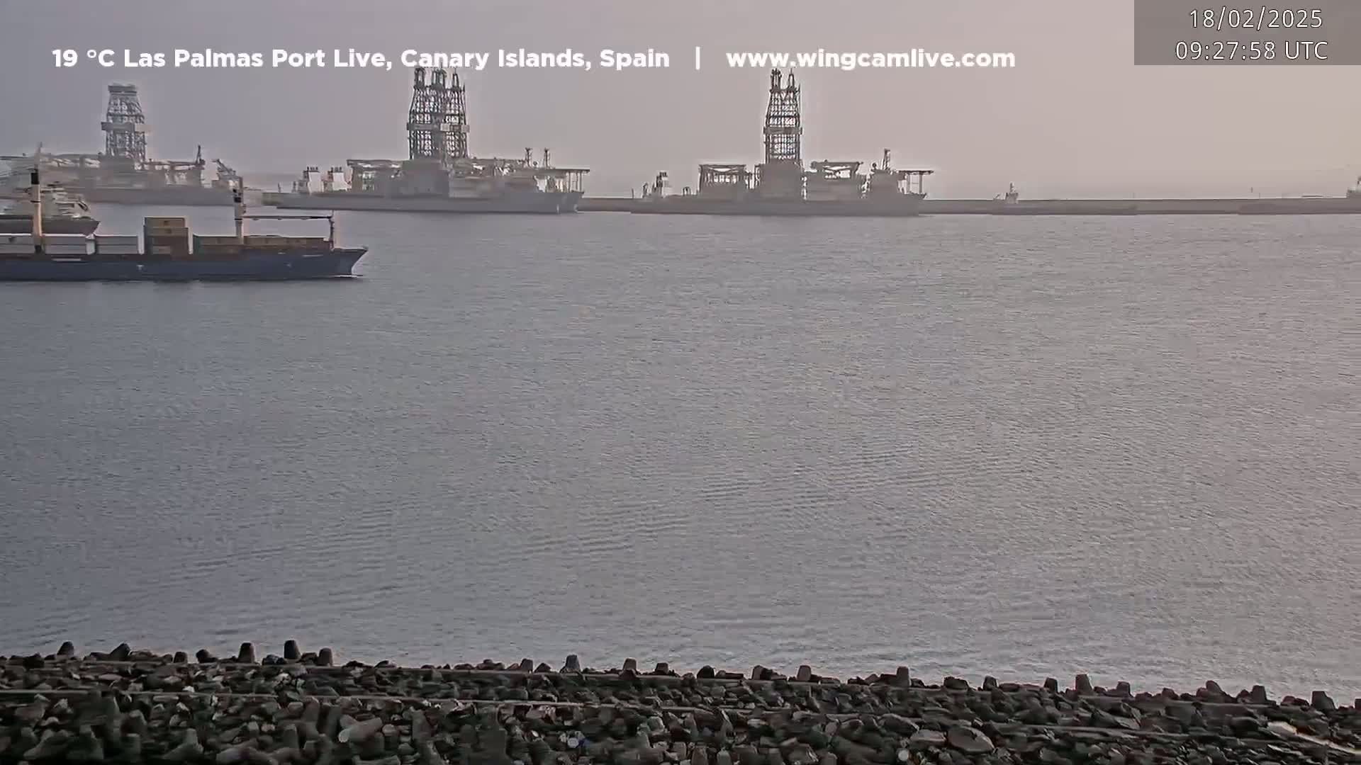 A container ship and several drilling rigs are visible in a hazy harbor.