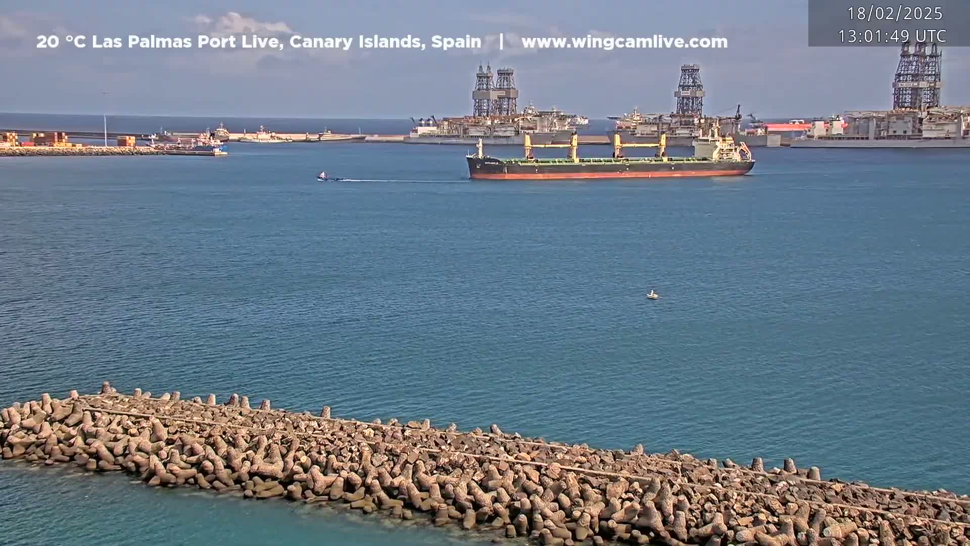 A large cargo ship is in the harbor near a breakwater on a calm, sunny day.