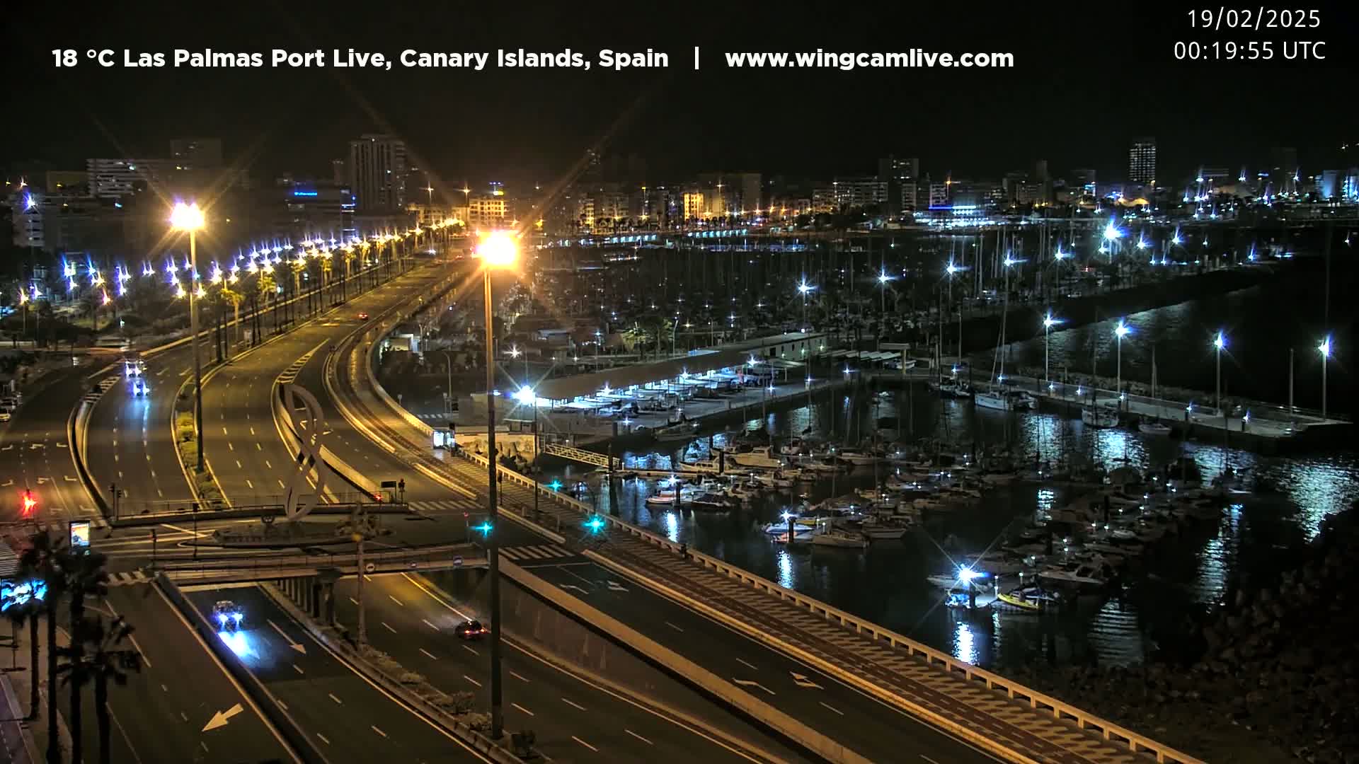 A nighttime aerial view shows a well-lit highway beside a marina filled with boats, all under a clear sky.