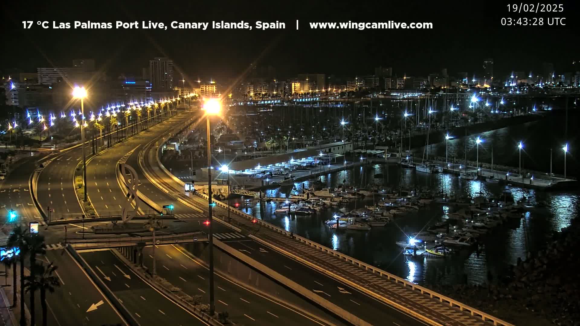 A nighttime view of a marina filled with boats, adjacent to a well-lit highway, under clear skies.