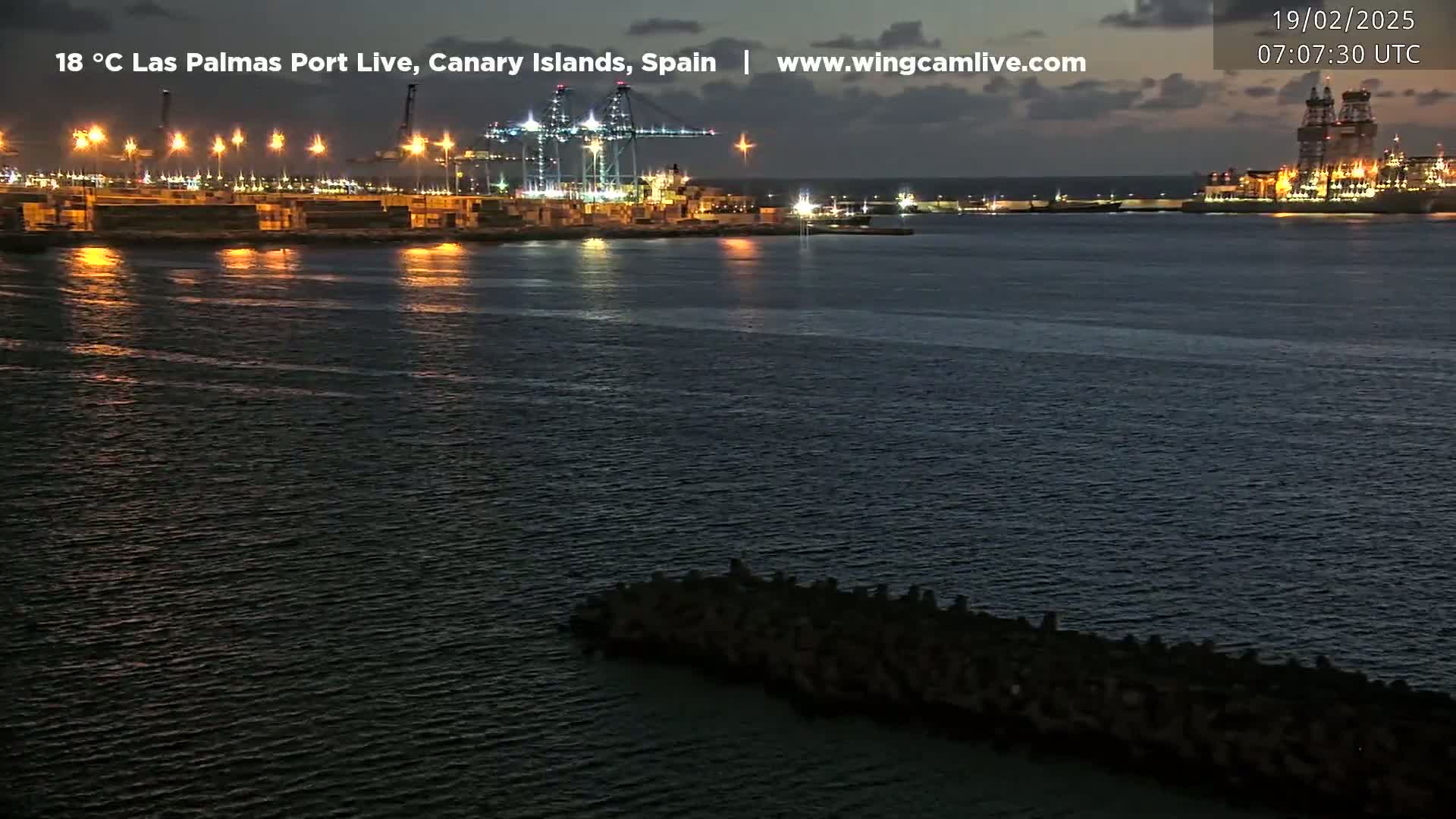 A nighttime view of a port with illuminated cranes and ships, calm water reflecting the lights, and a dark breakwater in the foreground.