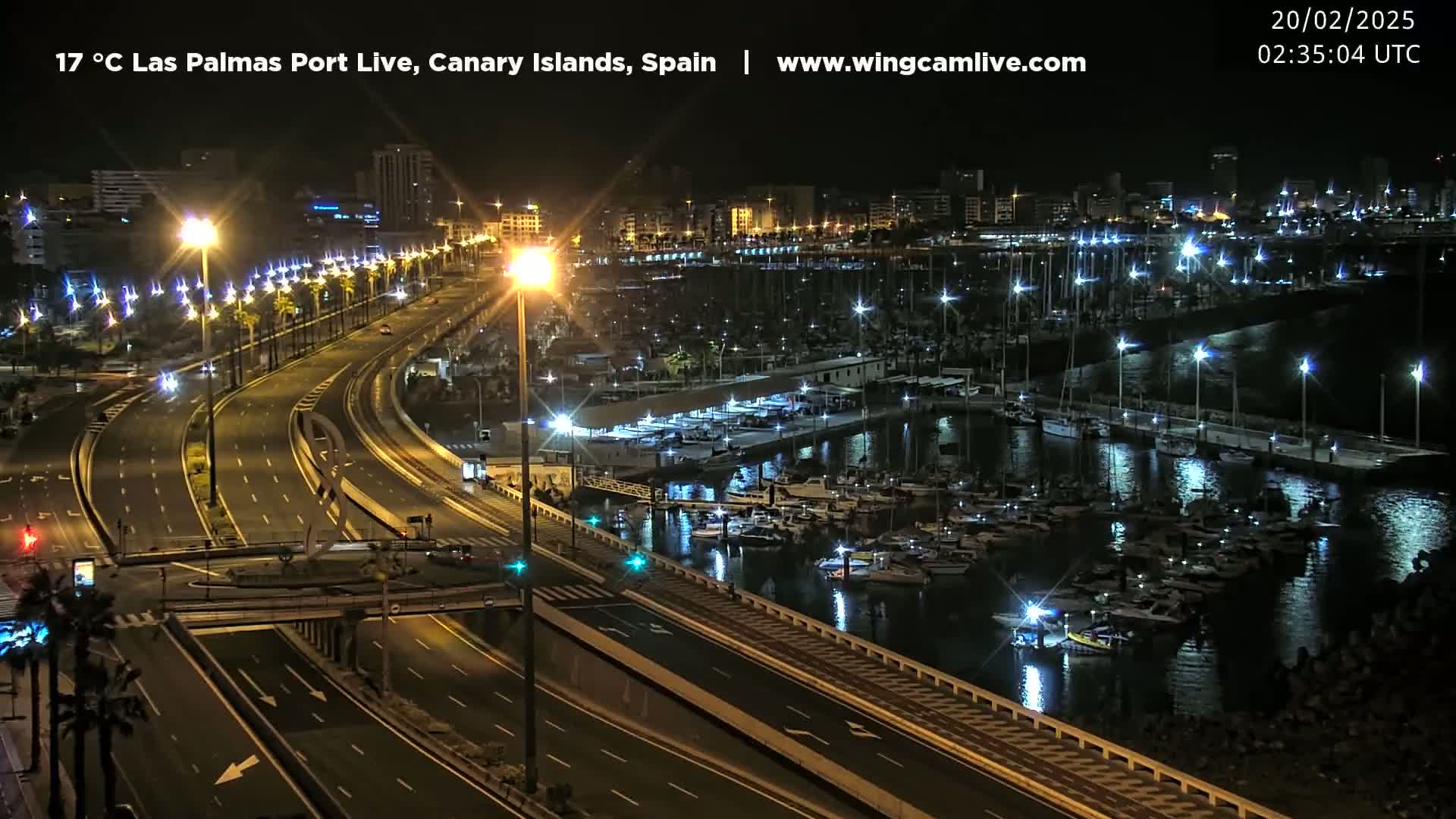 A nighttime view of a city harbor with many boats docked, a well-lit highway curving through the foreground, and a cityscape in the background.