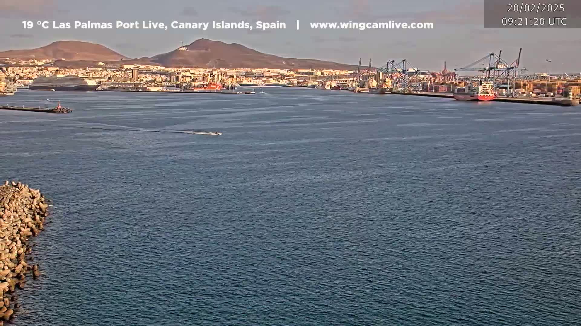 A calm body of water shows a port city with docked ships and cargo cranes under a clear sky.