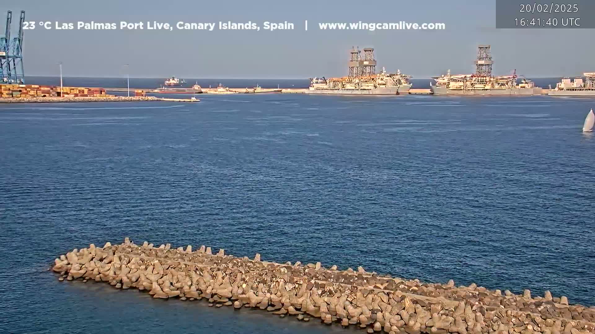 A calm, sunny day shows a harbor with several large ships docked, a breakwater in the foreground, and a sailboat in the distance.