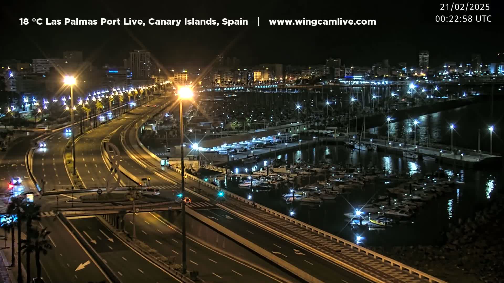 A nighttime view of a harbor filled with boats, next to a multi-lane highway with sparse traffic, under clear skies.