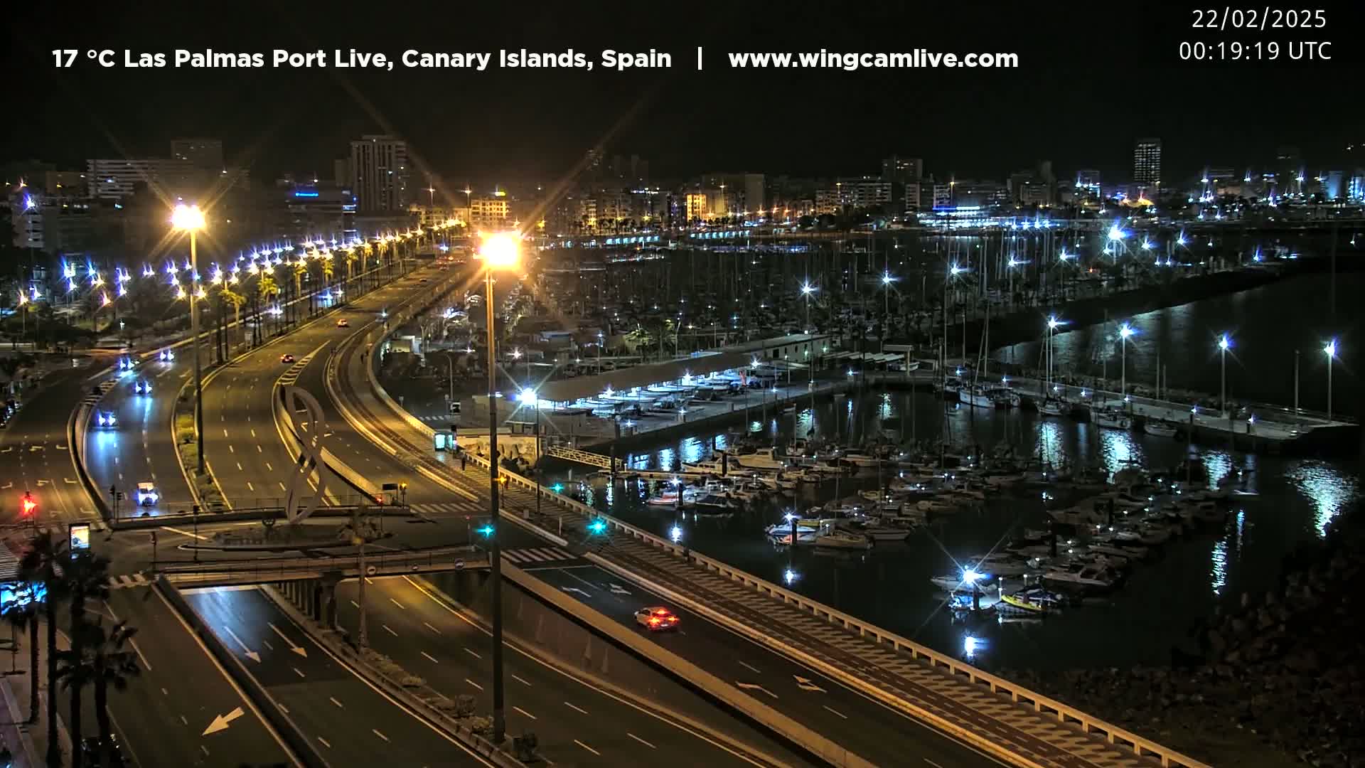A nighttime, well-lit view of a harbor filled with boats and a multi-lane highway next to it, with city lights in the background.