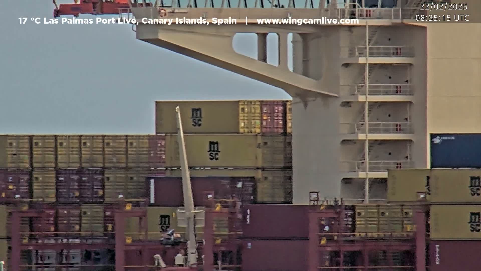 A container ship is being loaded with cargo containers at a port under a mostly clear sky.