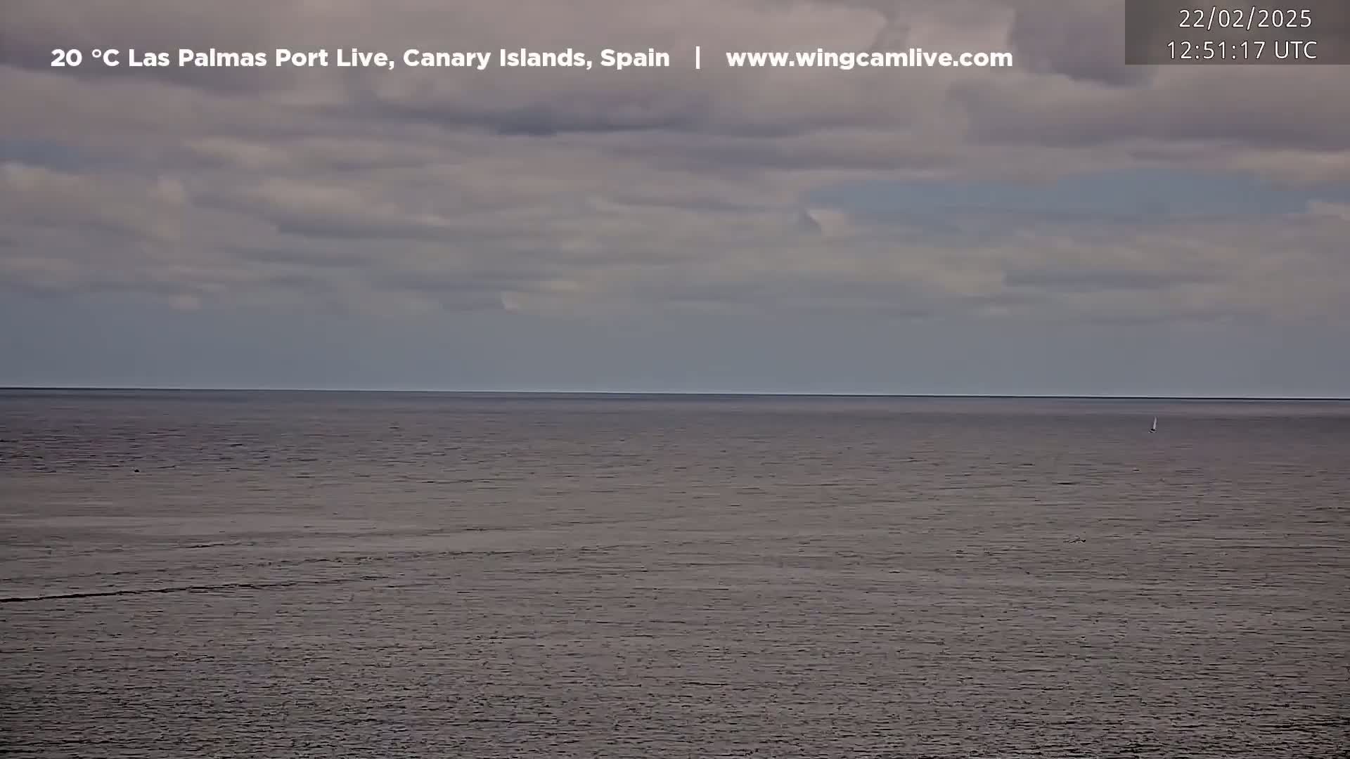A calm ocean under a mostly cloudy sky is seen from a distance, with a single sailboat visible on the horizon.