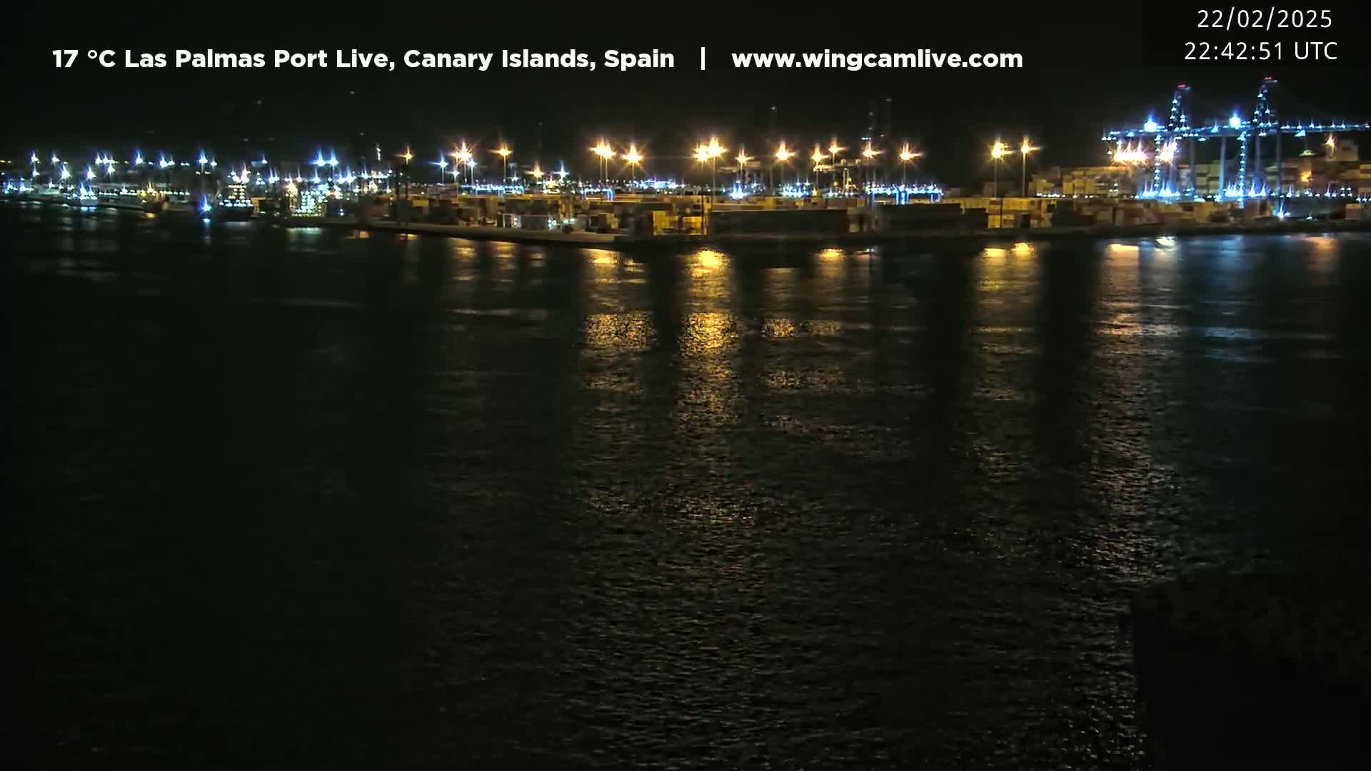A nighttime view of a brightly lit port with numerous containers and cranes reflected in calm water.