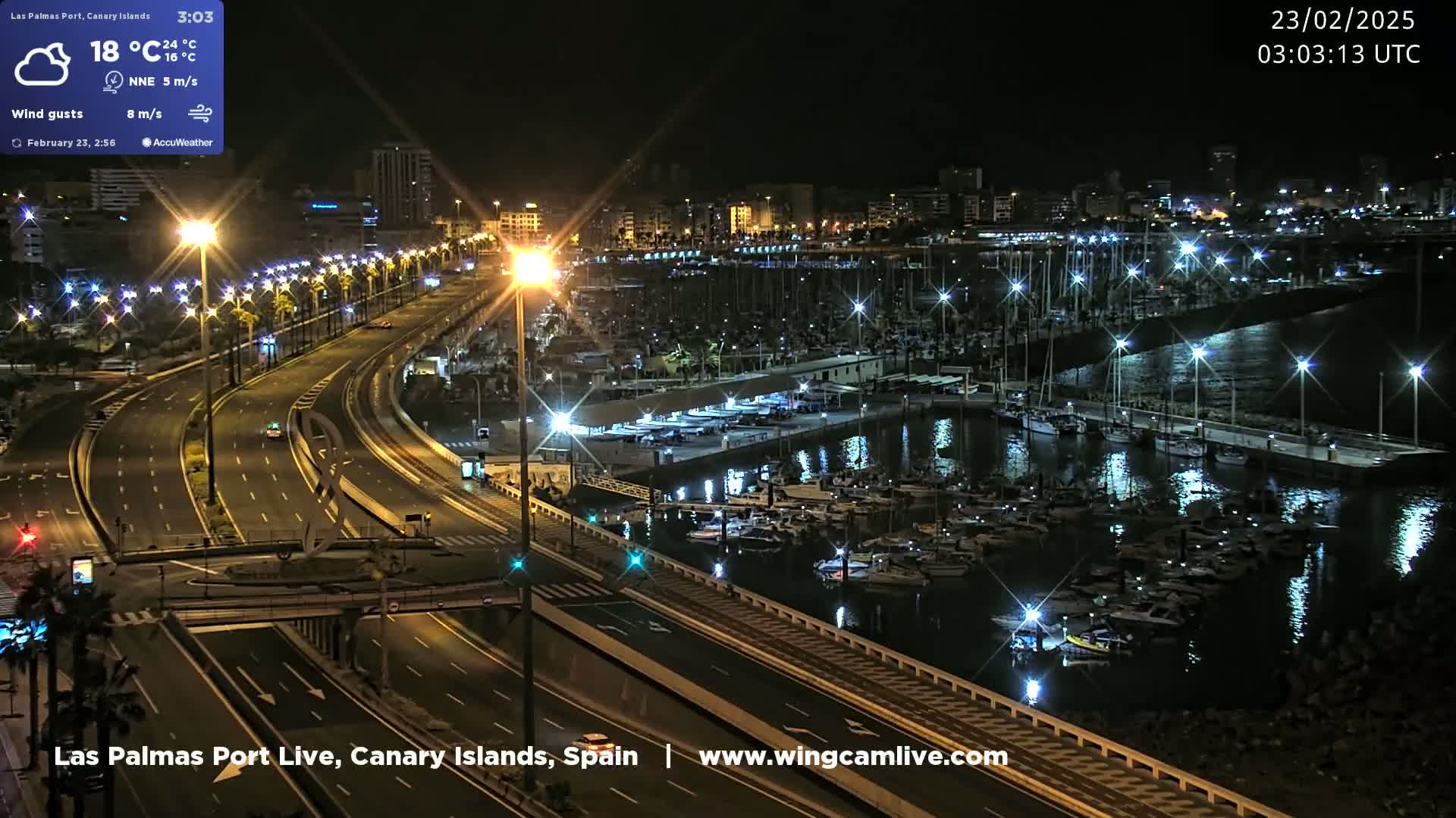 A nighttime aerial view of a harbor filled with boats, a multi-lane highway with streetlights, and a city skyline in the background.