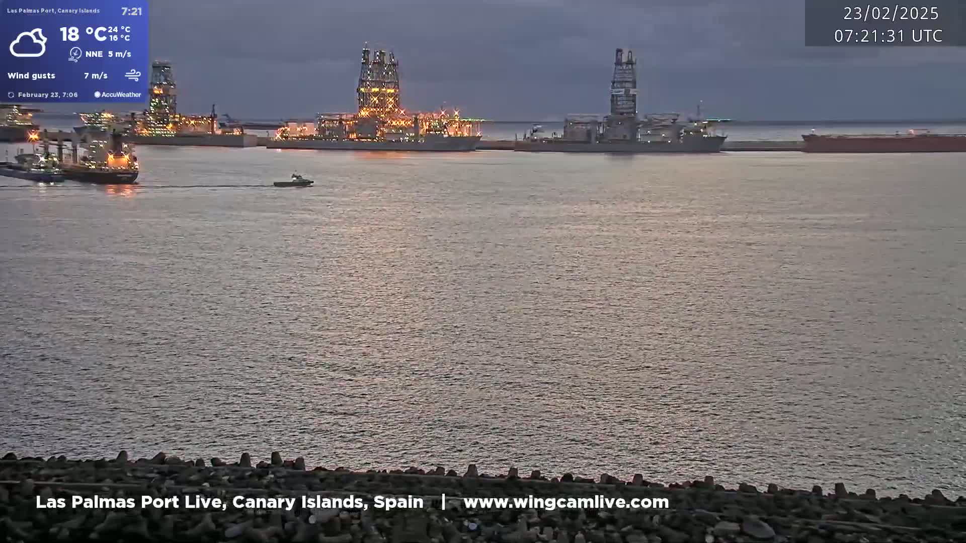 Several ships, including at least two large drilling platforms, are moored in a harbor at dusk under a mostly cloudy sky.