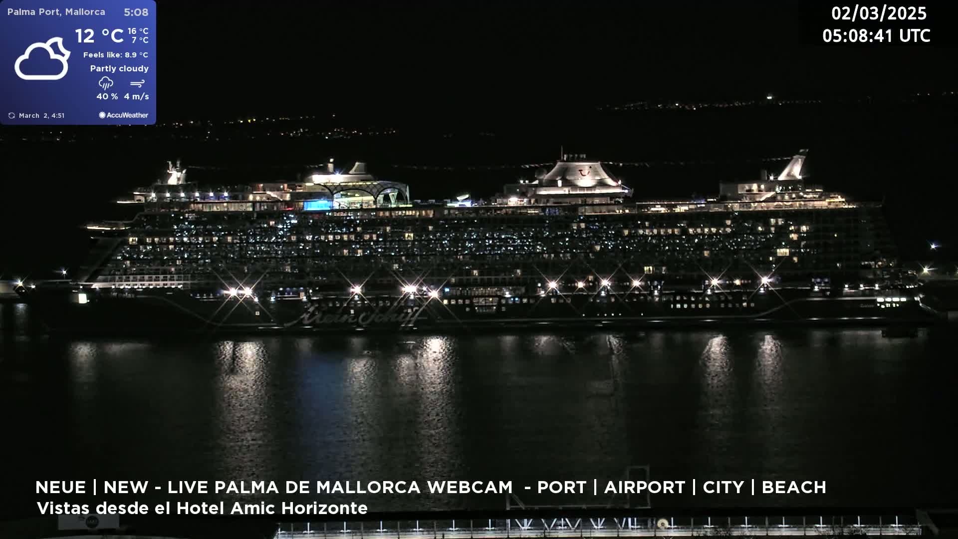 A large, brightly lit cruise ship is docked at a port at night under partly cloudy skies.
