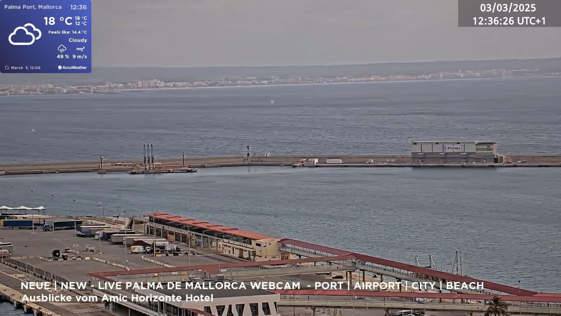 A cloudy day at a harbor shows a body of water, a distant shoreline, and various port buildings and structures, including a large building with a visible logo.