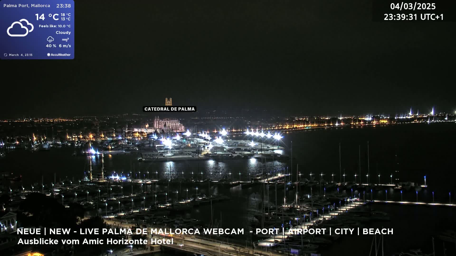 A nighttime, cloudy view of a harbor filled with boats, lit by many lights, with a city skyline and a large cathedral in the background.