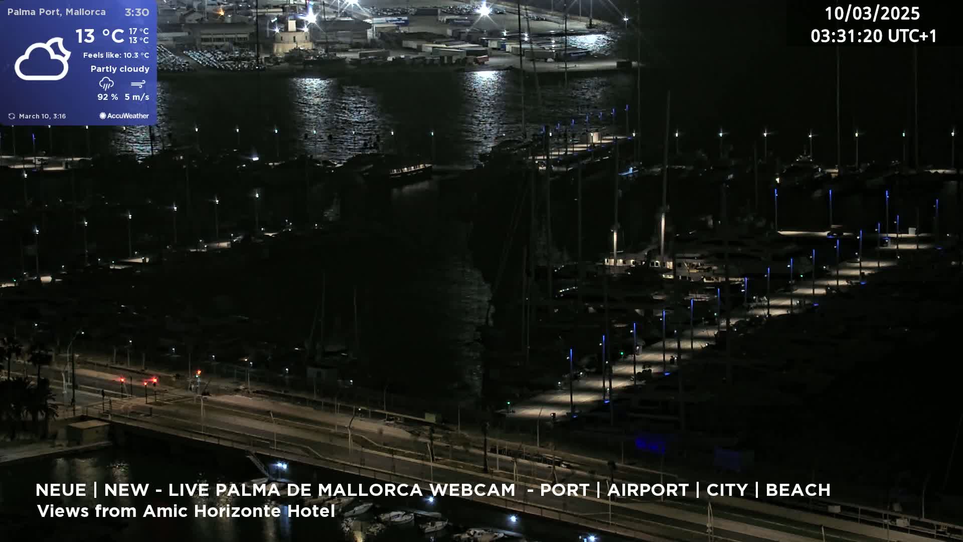 A nighttime, partly cloudy view of a marina filled with boats, illuminated by lights, next to a road with sparse traffic.