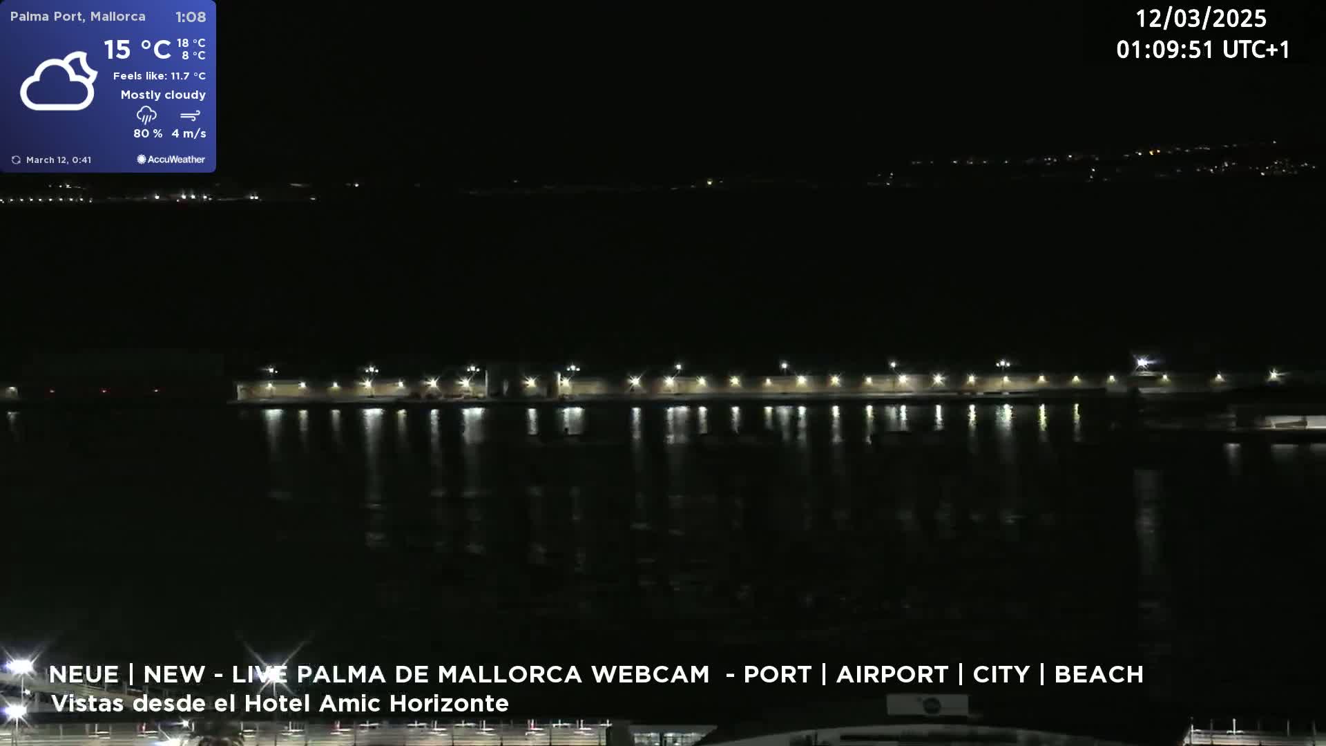 A nighttime view of a waterfront area with lights reflecting on calm water under a mostly cloudy sky.