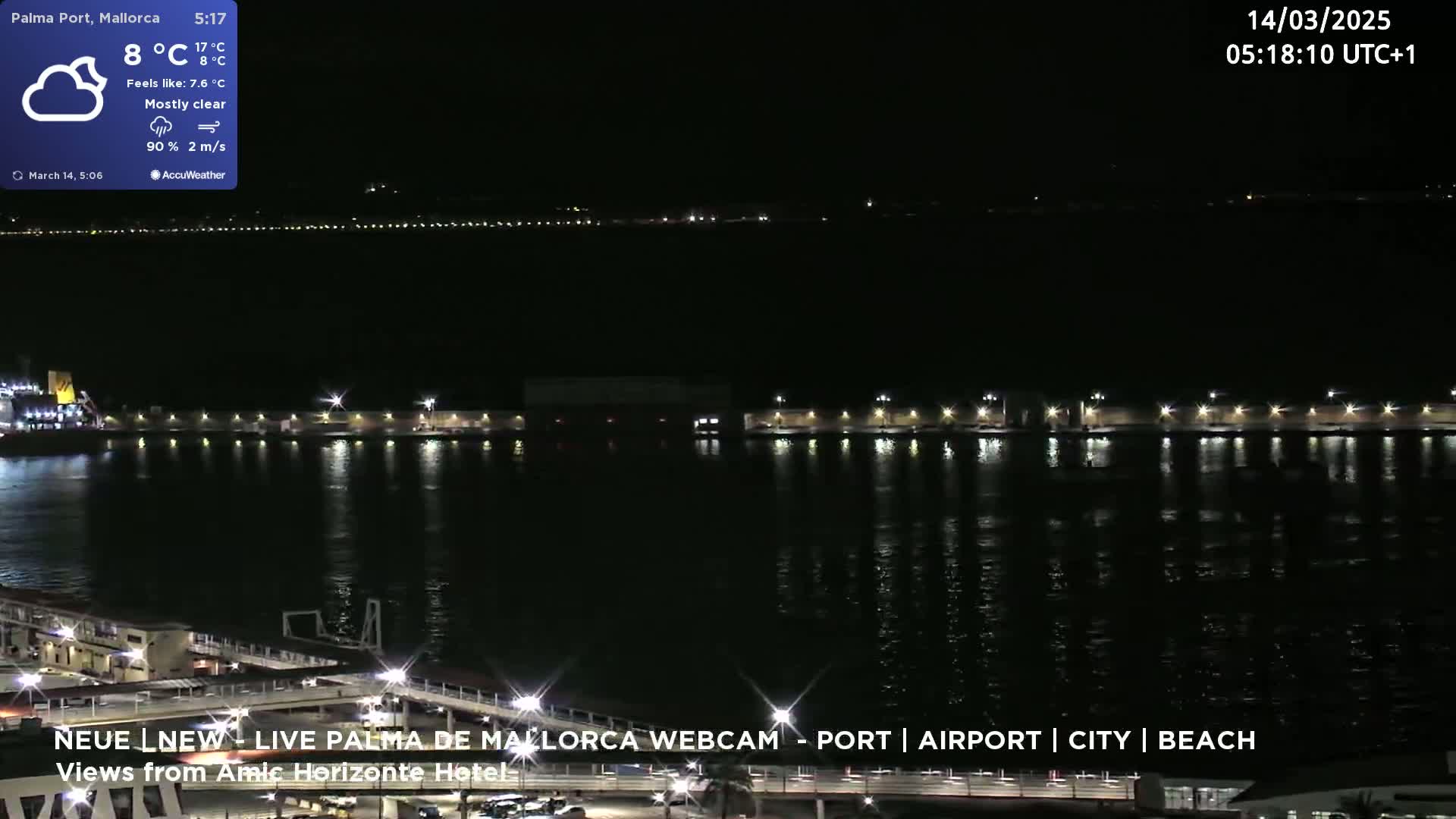 A nighttime harbor scene with calm water reflecting the lights of a pier and distant city lights under mostly clear skies.