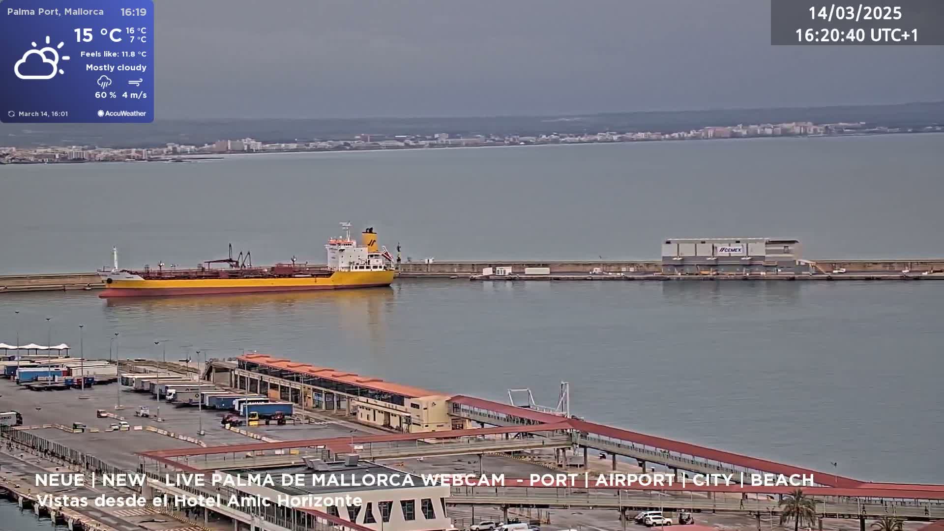 A mostly cloudy day shows a yellow and red tanker ship docked at a port with several vehicles parked nearby and a city visible across the water.