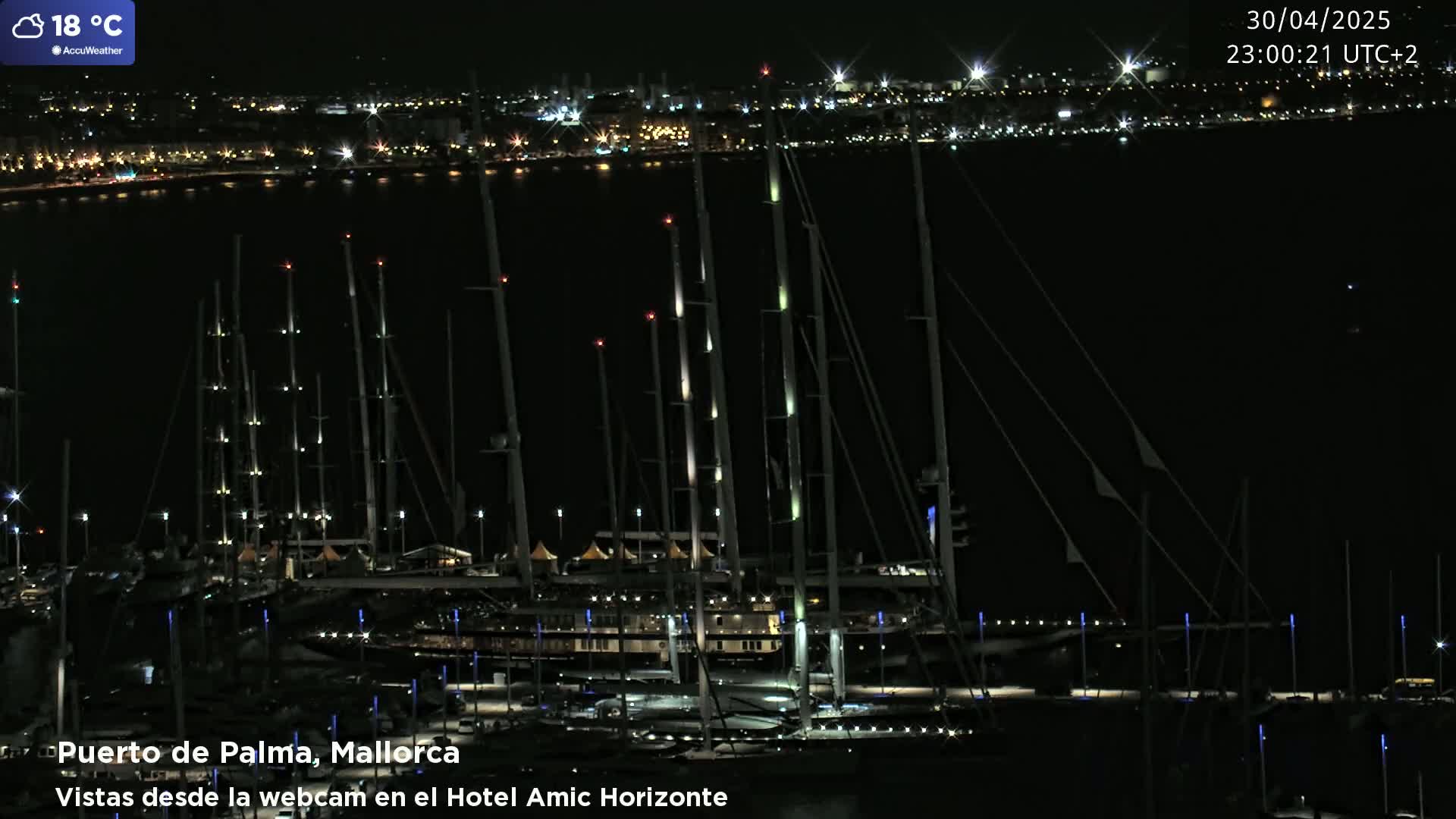 Numerous sailboats are docked at a marina at night, illuminated by lights, with a city skyline visible in the distance.