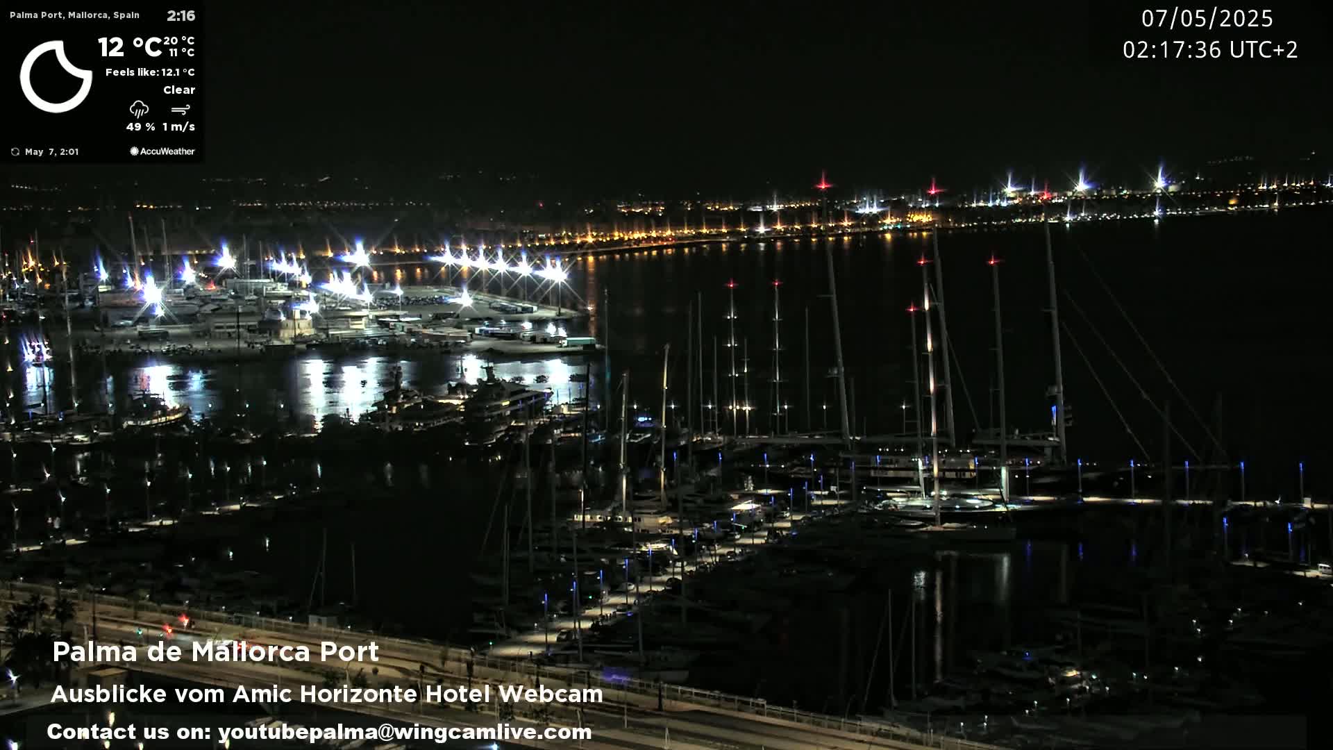 A nighttime view of a marina filled with boats, illuminated by lights, with a city skyline visible in the distance under clear skies.
