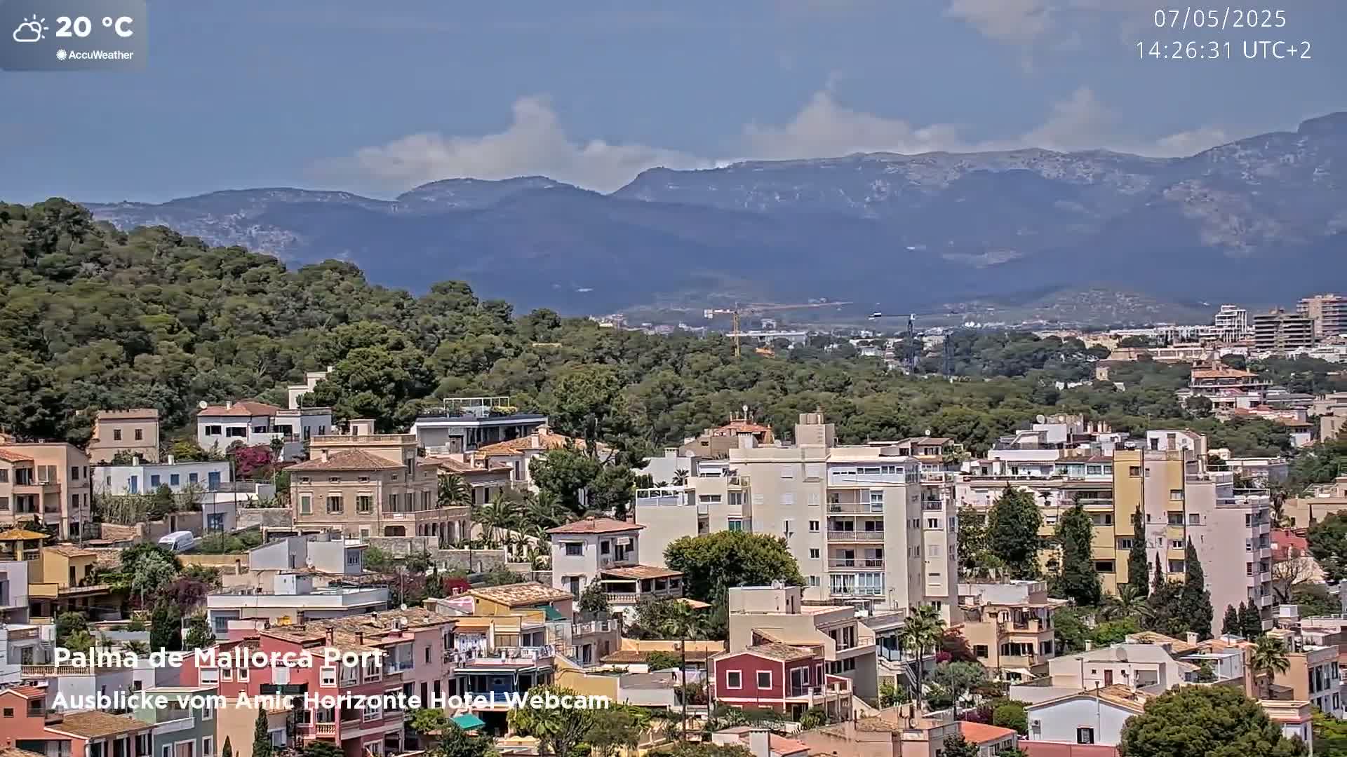 A sunny day shows a panoramic view of a town nestled amongst green hills and mountains.
