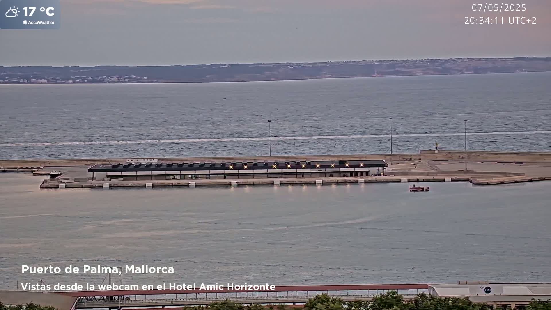 A harbor scene at dusk shows a long, low building on a pier extending into calm water, with a distant shoreline visible under a cloudy sky.