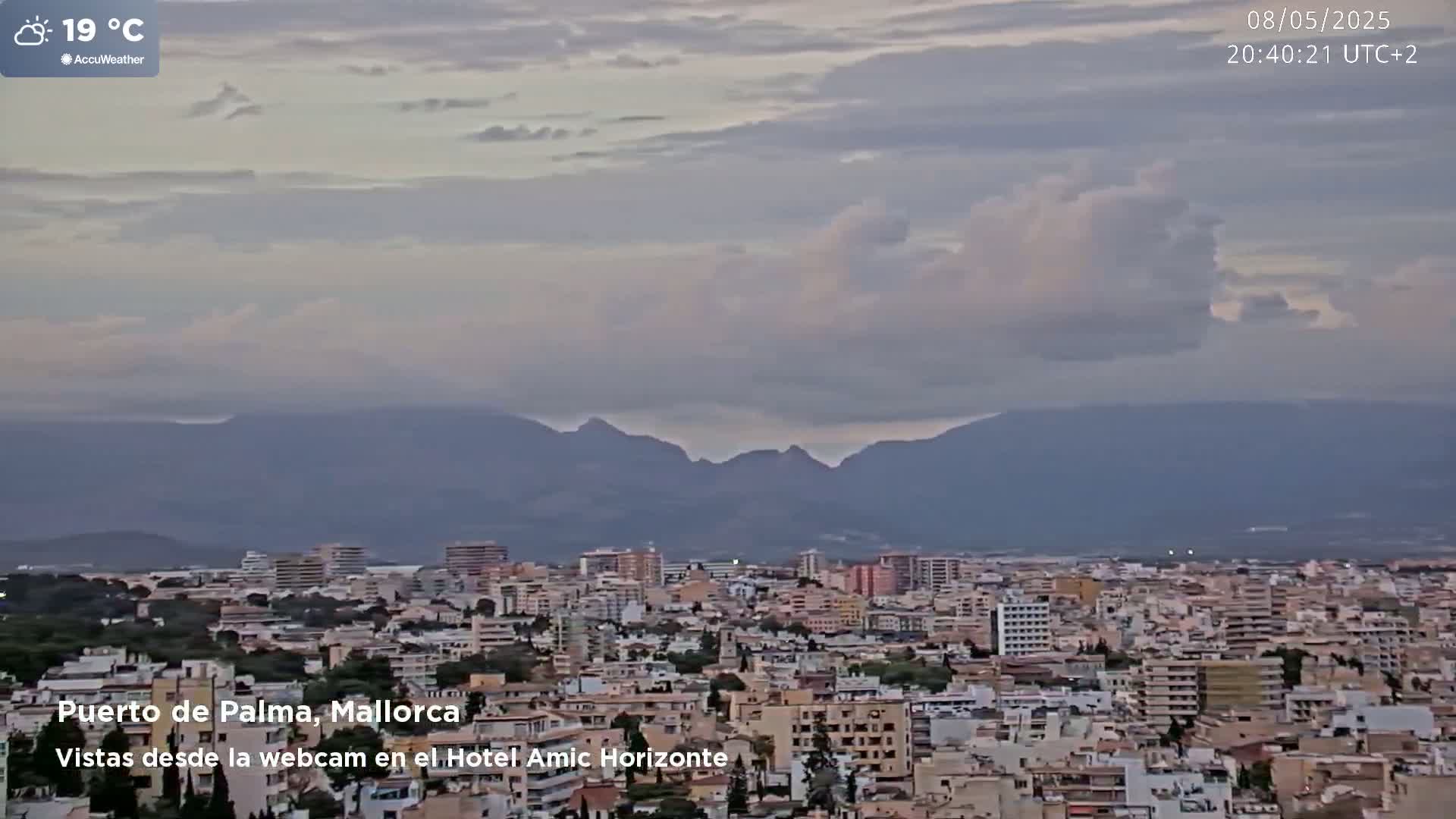 A sprawling city, under a partly cloudy sky at 19°C, is nestled against a range of distant mountains.