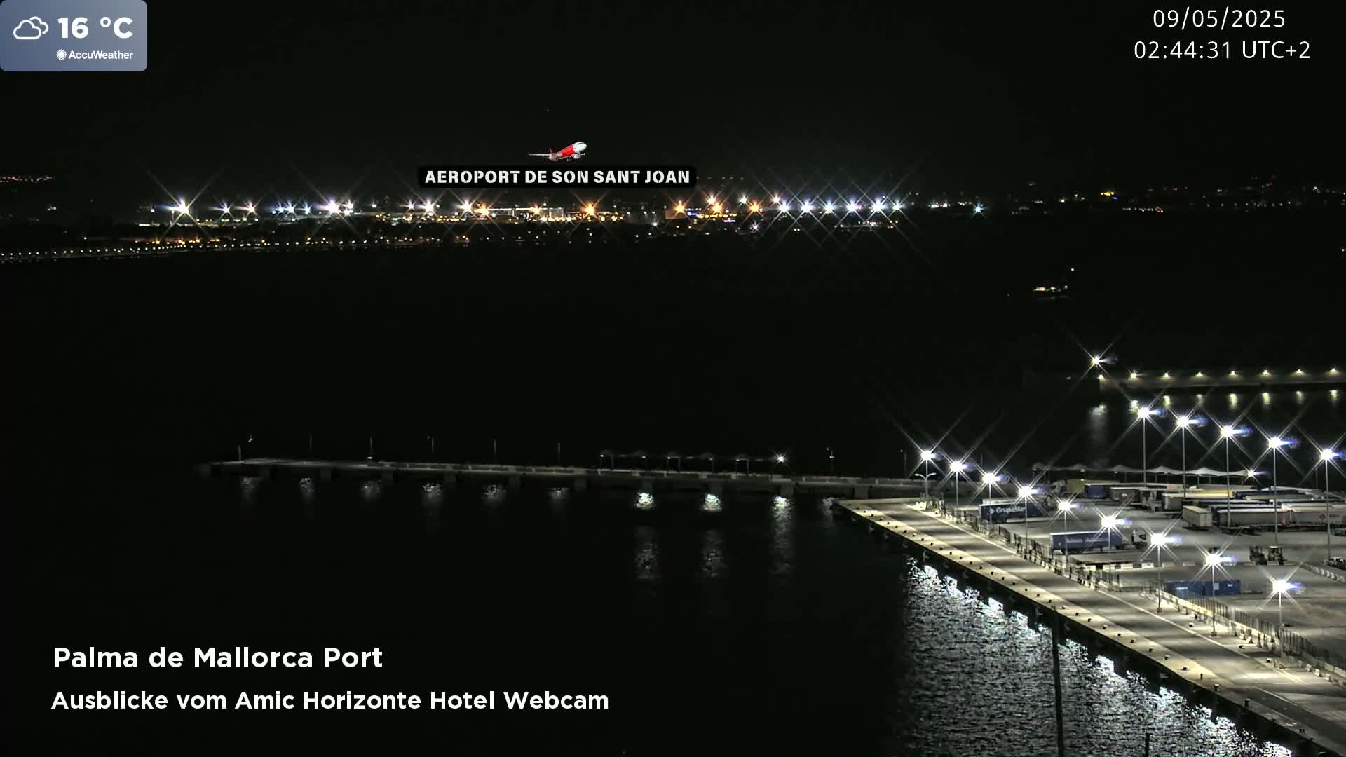 A nighttime view of a brightly lit port with an airplane flying over a distant, also illuminated, airport.