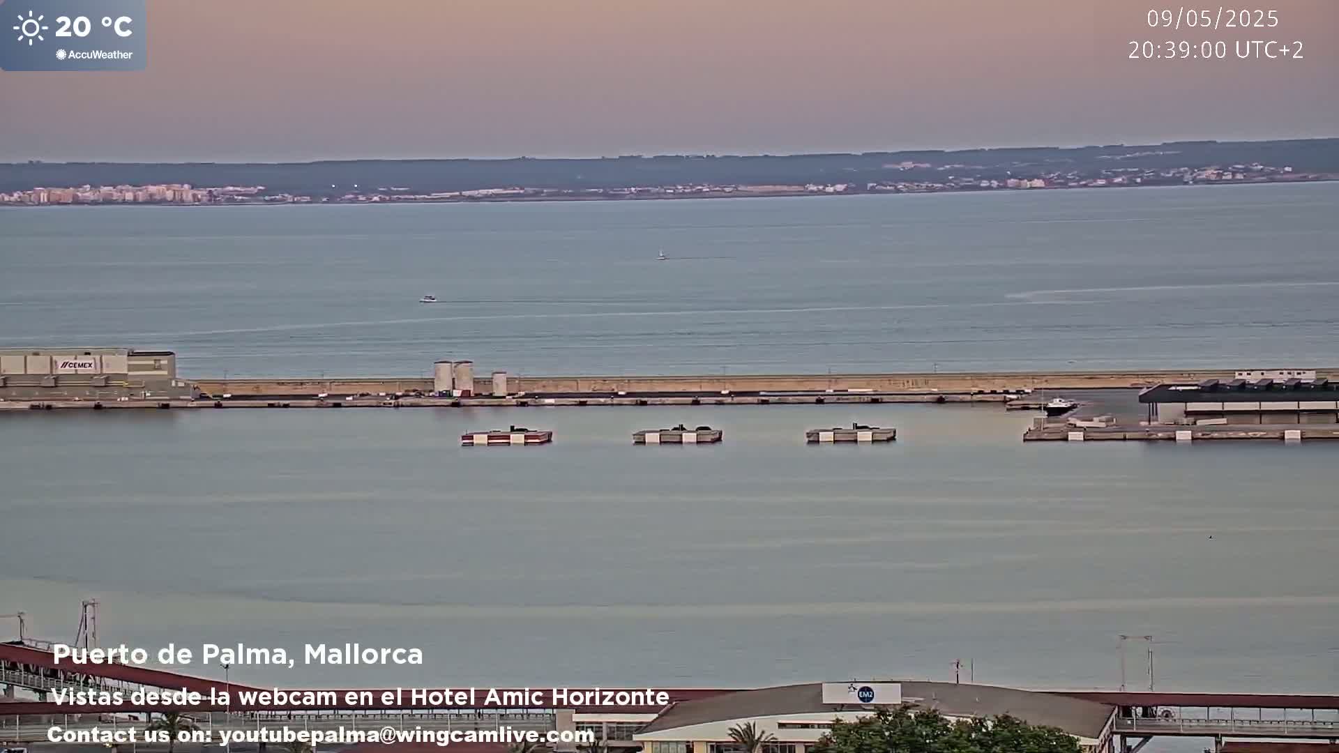 A calm harbor with several moored barges is seen at dusk, with a city skyline visible in the distance across the water.