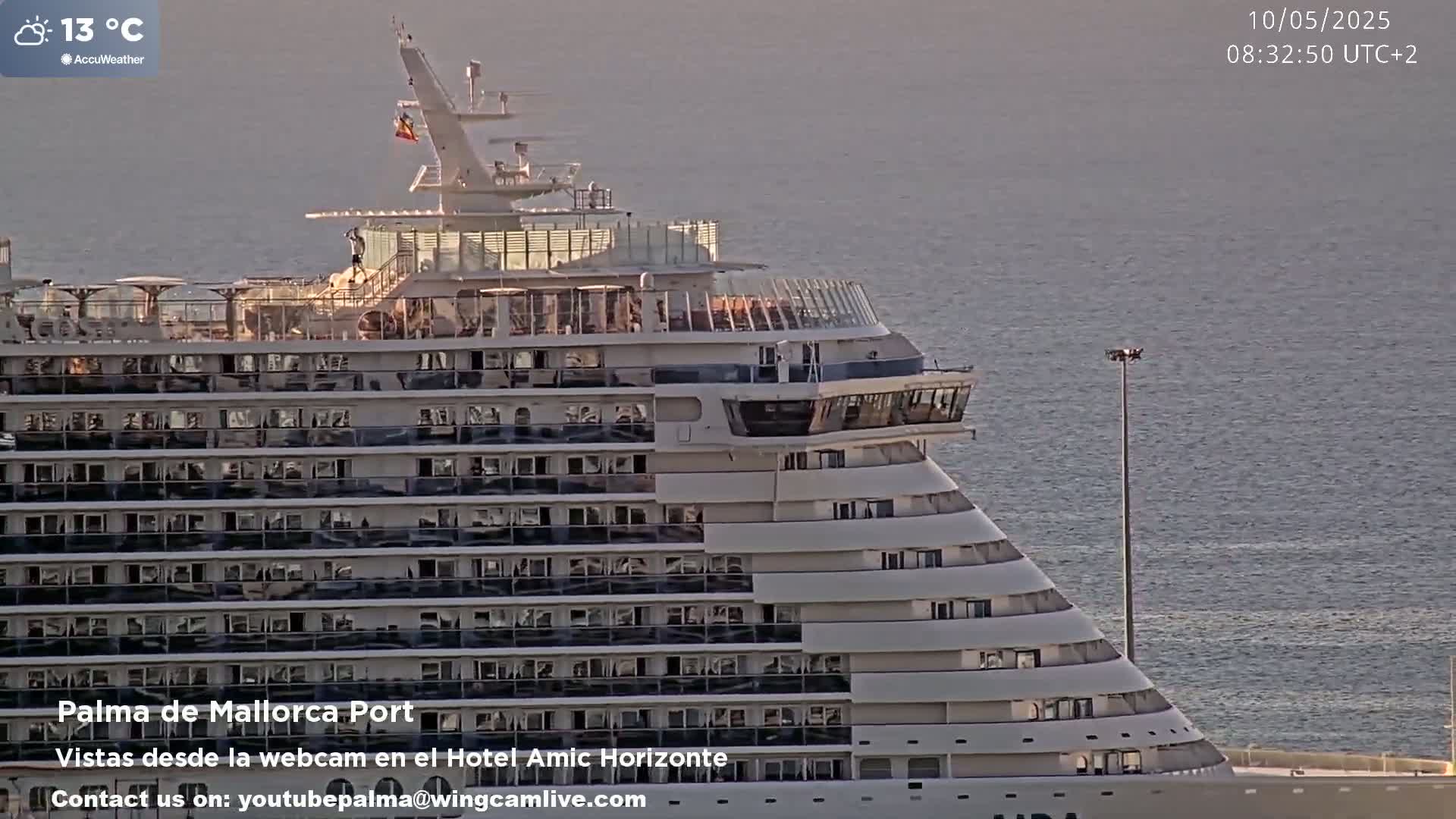 A large cruise ship is docked at a port on a calm, sunny day with a temperature of 13°C.