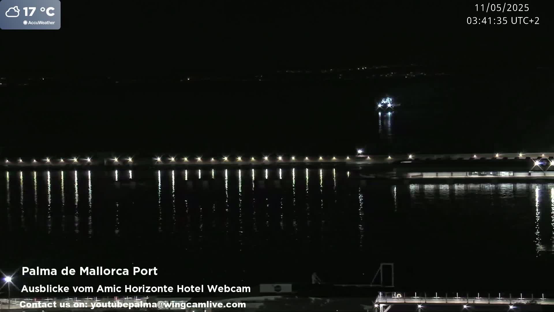 A nighttime view of a harbor with a lighted pier reflecting in calm water, and a distant vessel.