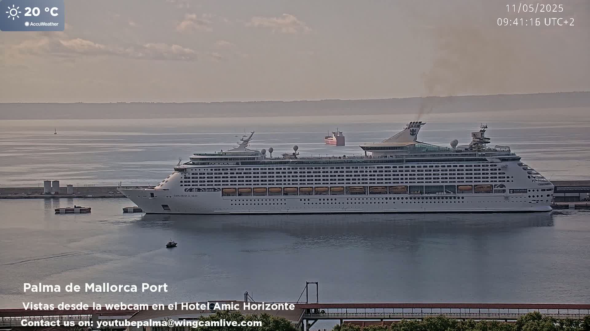 A large cruise ship is docked in a calm harbor on a hazy, sunny day with a temperature of 20°C.