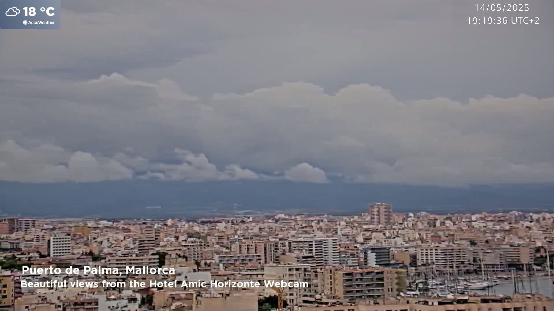 A cloudy, 18°C cityscape view shows a harbor filled with boats and many buildings extending to distant mountains.