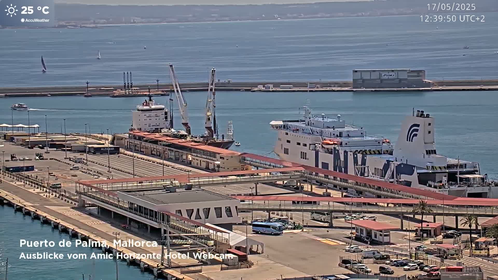 A sunny day at a port shows a large ferry docked at a terminal, alongside a cargo ship and several cranes, with cars and buses parked nearby.