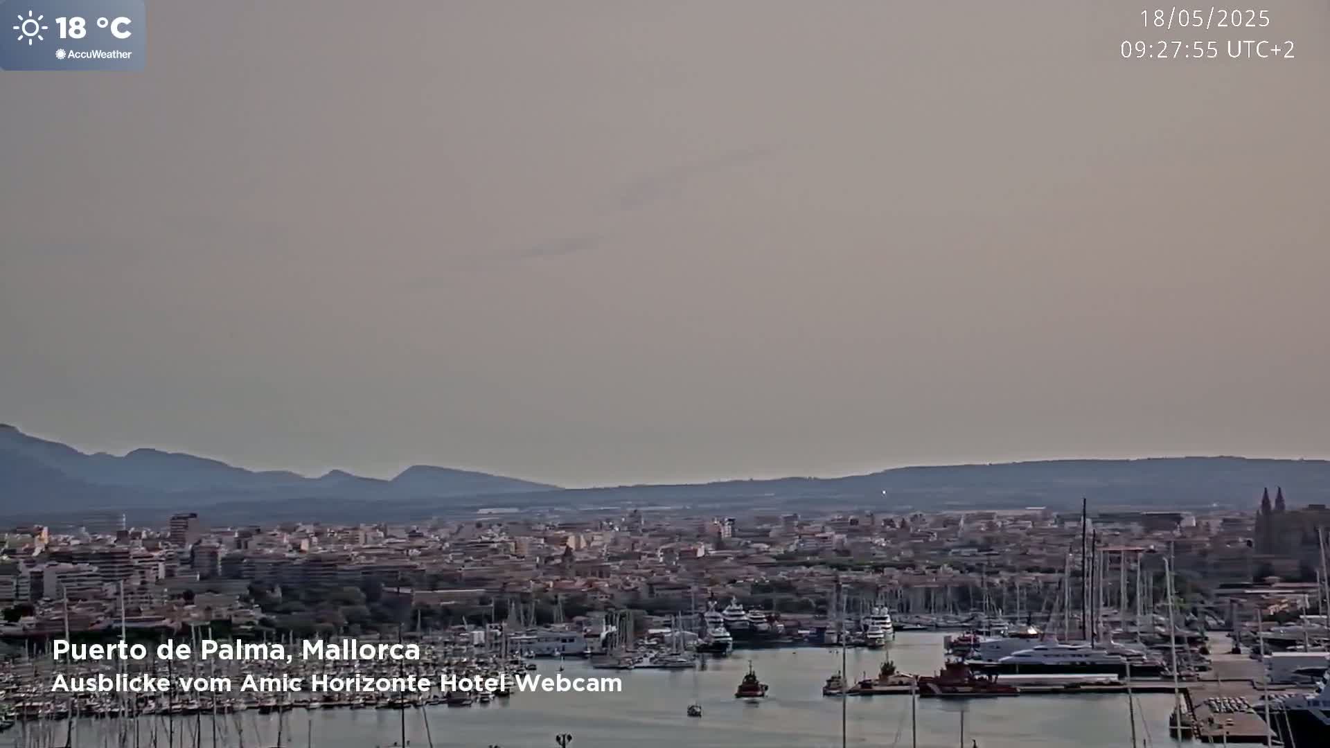 A hazy, daytime view shows a harbor filled with boats in front of a city nestled against mountains.
