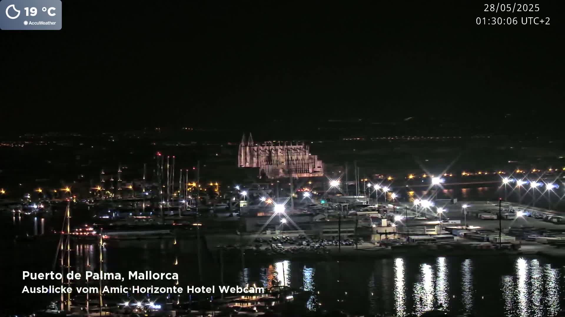 A nighttime harbor scene shows a large illuminated cathedral in the distance, boats docked along a waterfront, and bright lights reflecting on the water, with a temperature of 19 degrees Celsius.