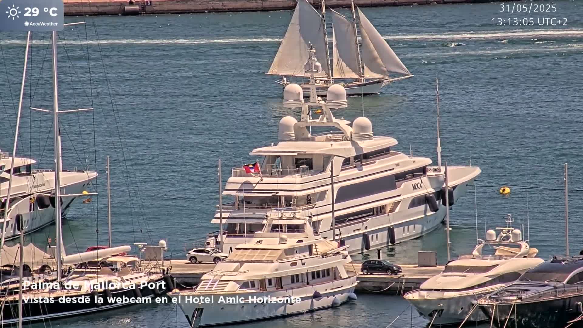 A large yacht is docked in a harbor on a sunny day, with several other boats and sailboats nearby.