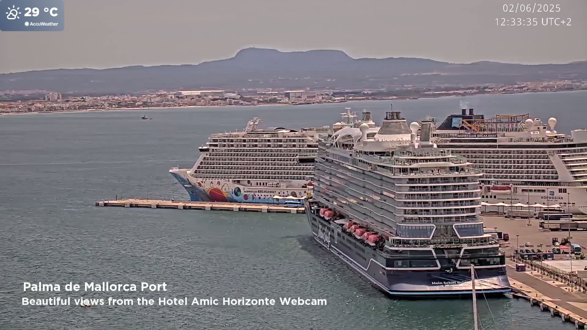 Two large cruise ships are docked at a port, with mountains visible in the distance on a sunny day.