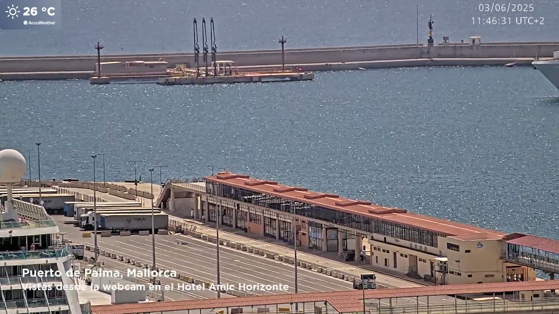 A cruise ship is docked at a port on a sunny day, with several trucks parked nearby and calm, blue water visible in the background.