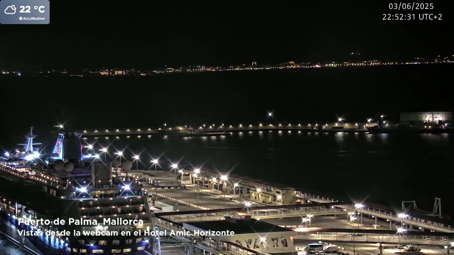 A nighttime view of a cruise ship docked at a brightly lit port, with a city skyline visible in the distance under a clear sky.