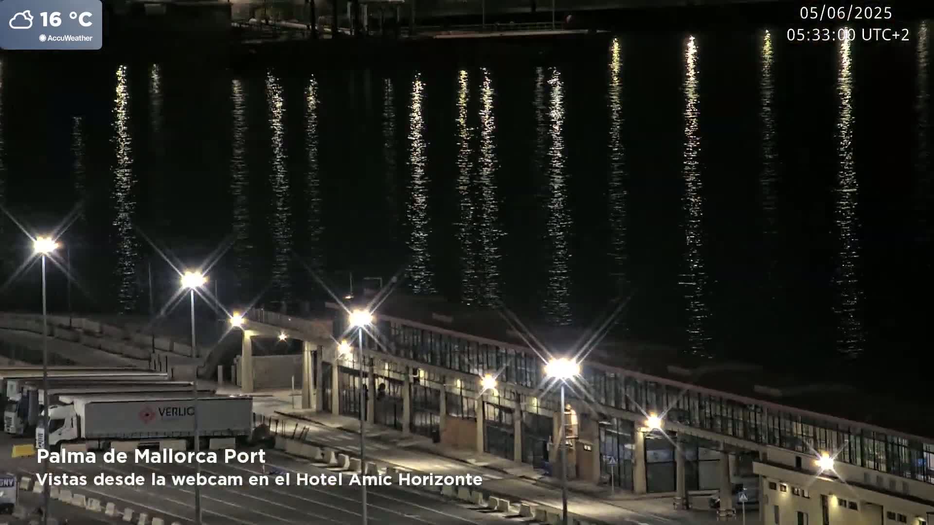 A nighttime view of a port with calm water reflecting lights from the shore, under a clear sky.