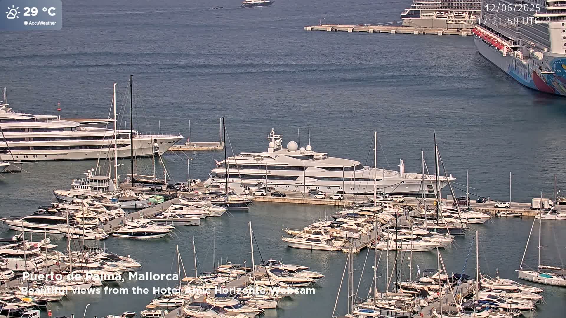 A large marina filled with numerous yachts and boats on a sunny day, with a large luxury yacht prominently featured in the center.