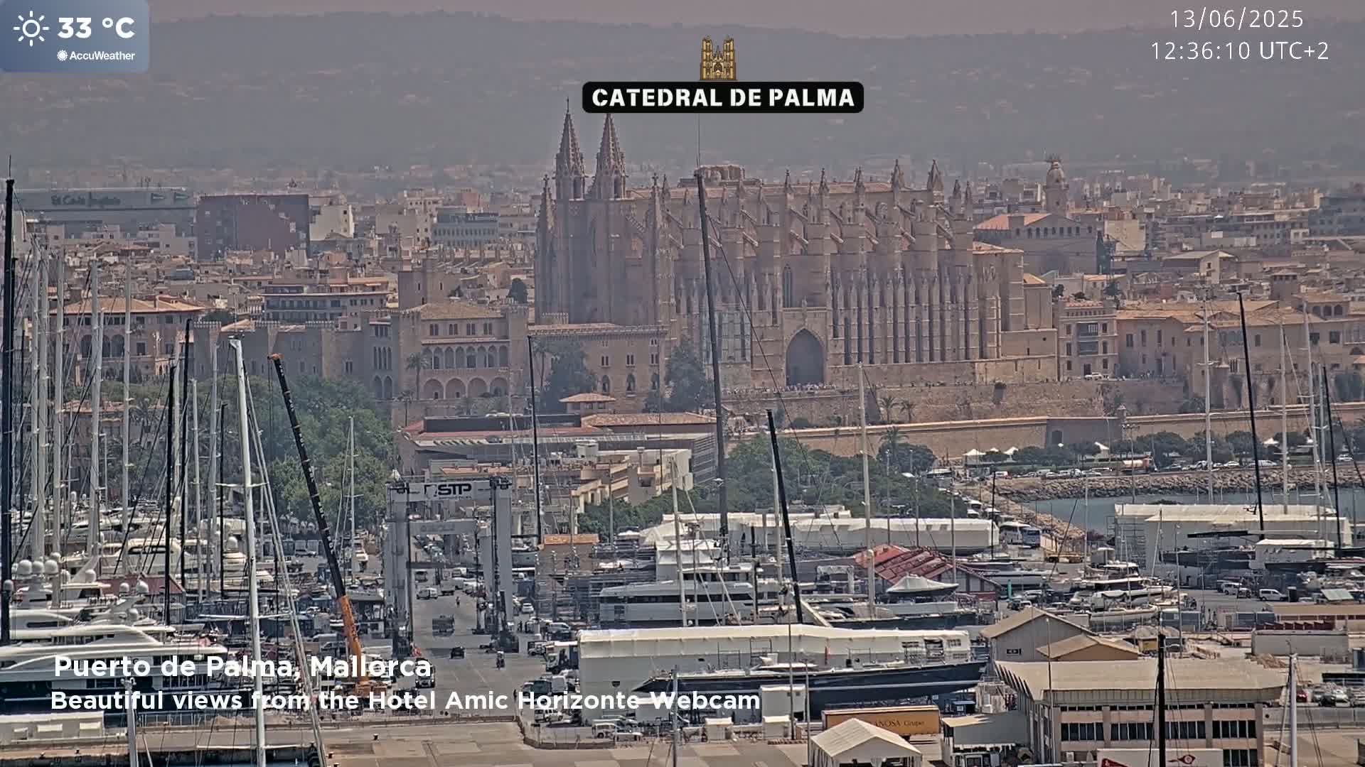 A hazy, sunny day at 33°C shows a harbor filled with sailboats and yachts in front of the Catedral de Palma in Palma de Mallorca.